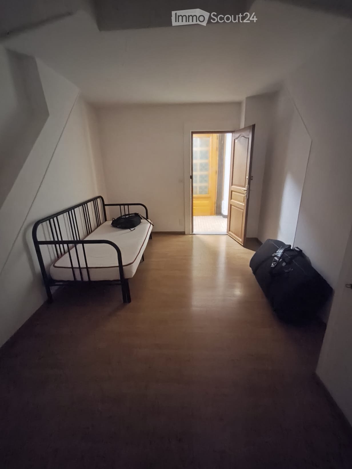 A small, furnished room with a single bed, a suitcase, and an open doorway leading to another room.