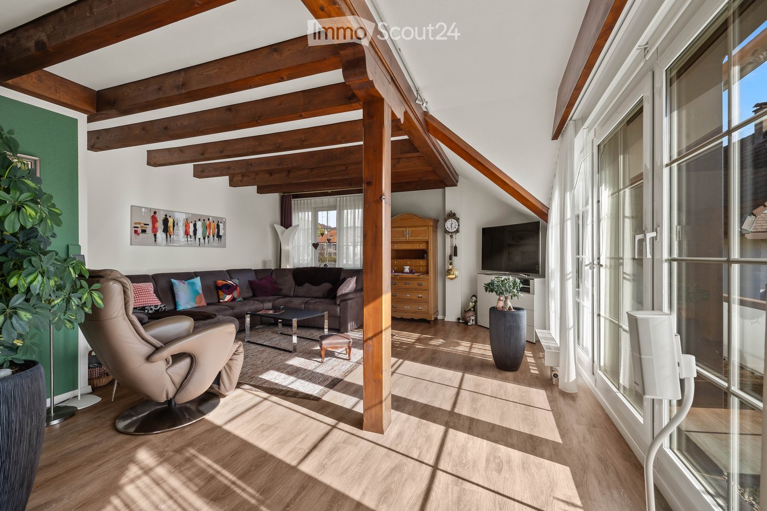 Bright living room with wooden beams, large windows, a dark brown sectional sofa, a coffee table, a leather armchair, a TV, and a wooden cabinet.