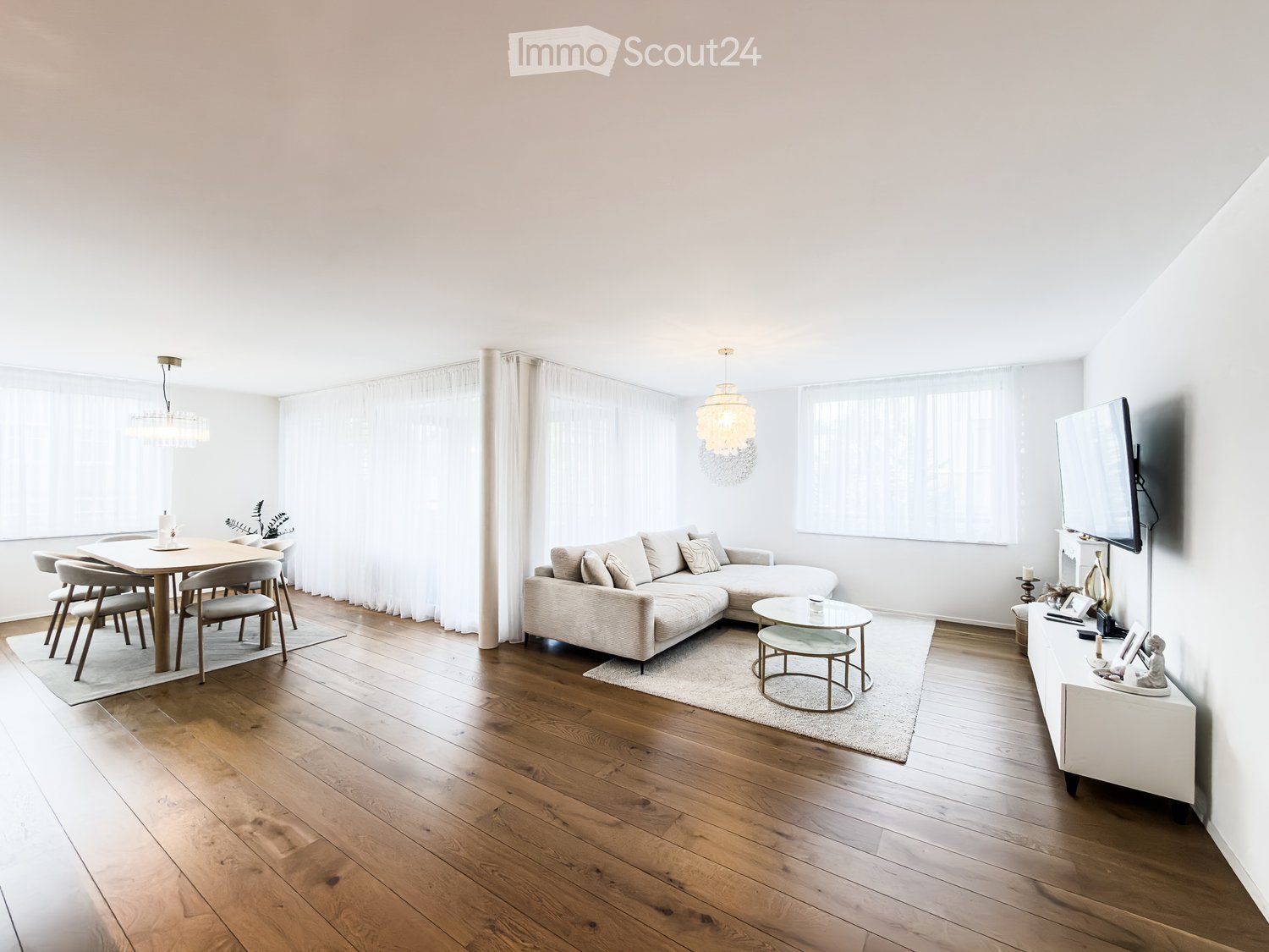 Bright, open living and dining area with wooden flooring, large windows, a sectional sofa, coffee table, dining table with chairs, and a TV on a stand.