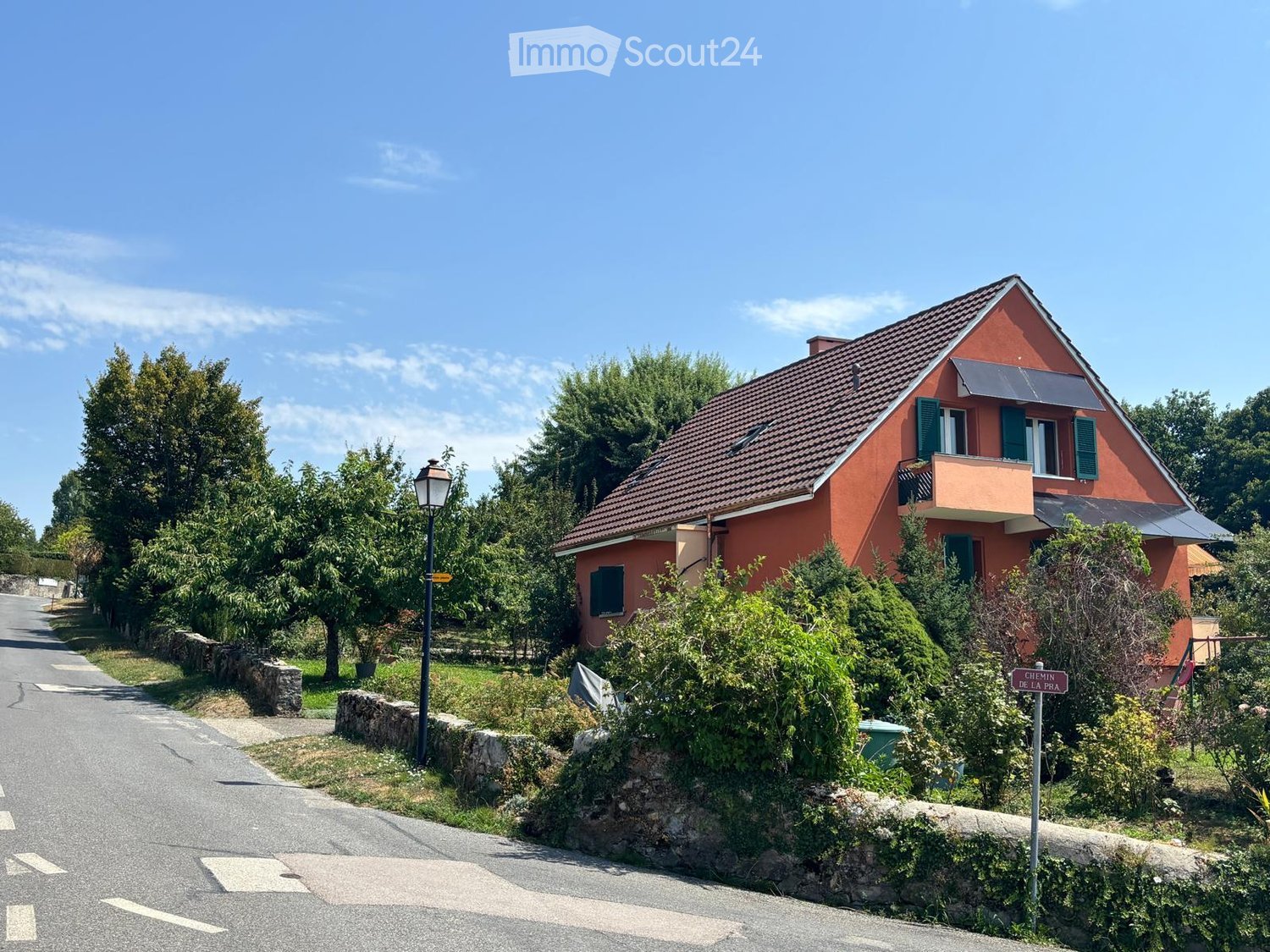 A two-story house with a red-tiled roof and green shutters, surrounded by trees and greenery, with a solar panel on the roof and a small balcony on the upper floor.