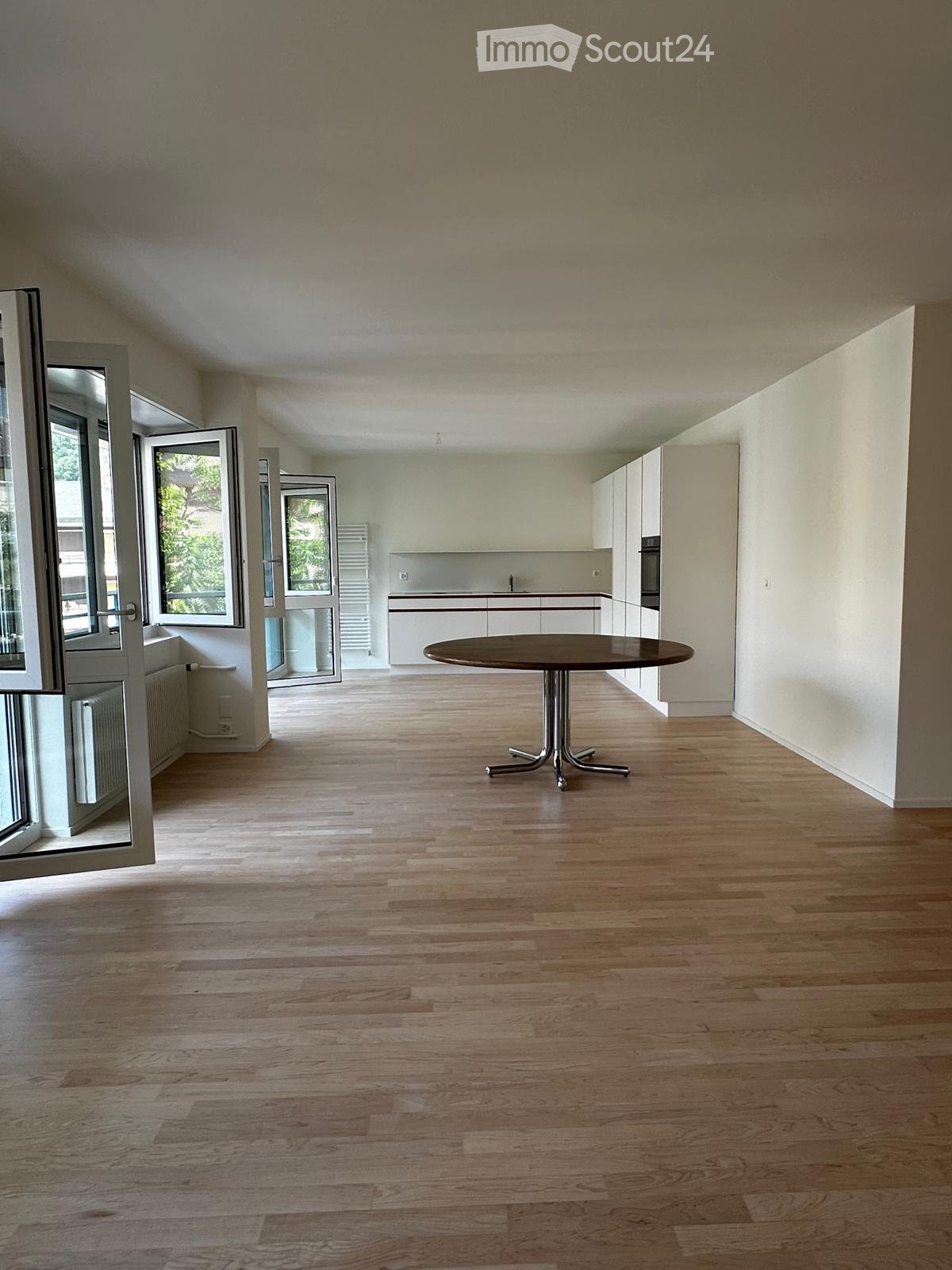 Open-plan space with light wooden flooring, white walls, and a single round table near the kitchen area. Large windows and sliding doors provide natural light and access to the outdoors.