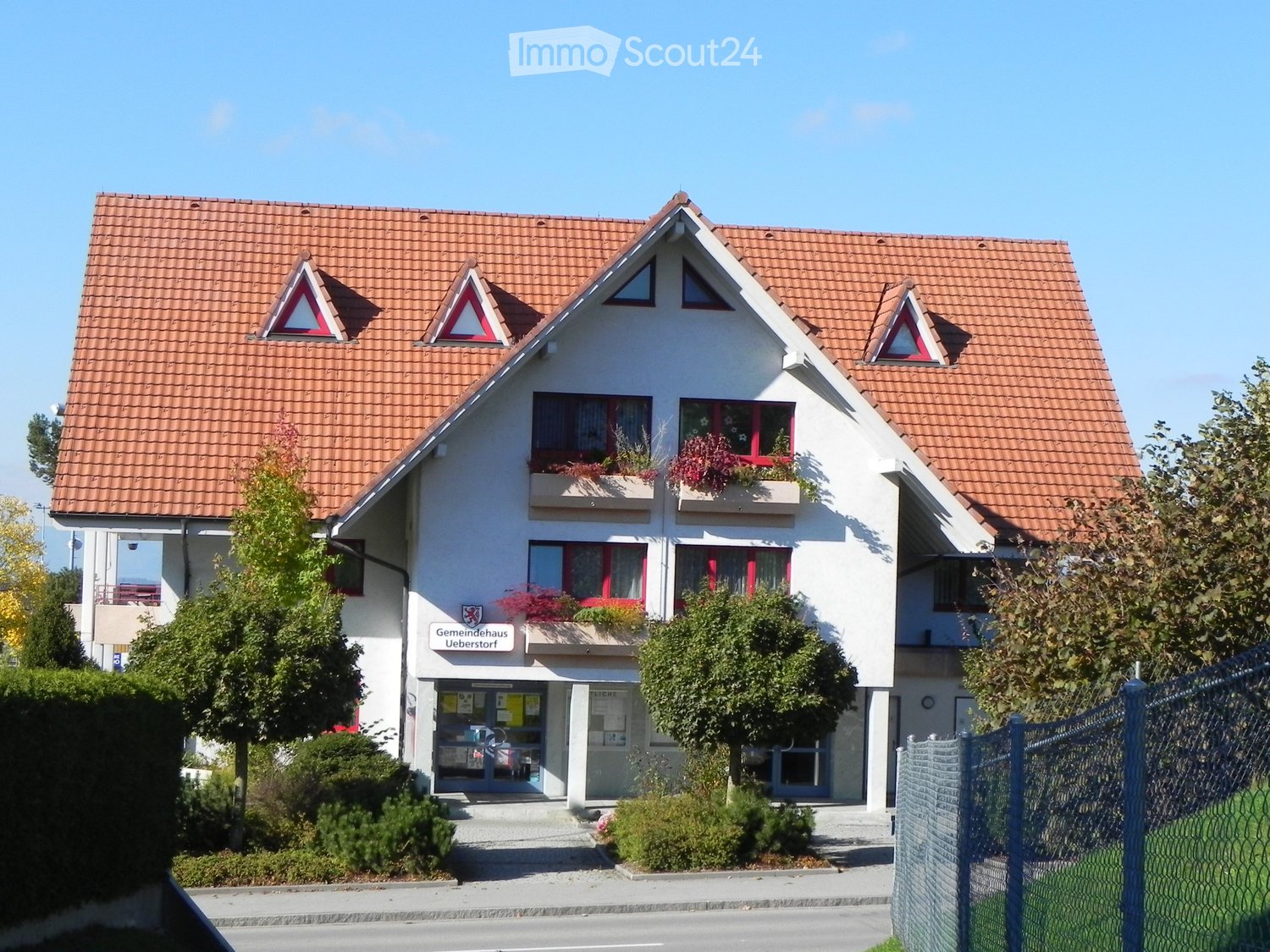 Two-story building with a red-tiled roof, white walls, and red-framed windows. Features a sign reading "Gemeindehaus Ueberstorf" and a fenced area in front.