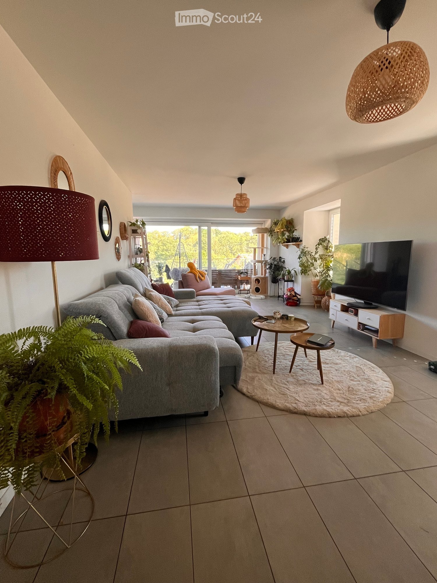 Modern living room with grey sectional sofa, coffee table, small round table, TV, and decorative lamps. Features large windows with a view of greenery outside.