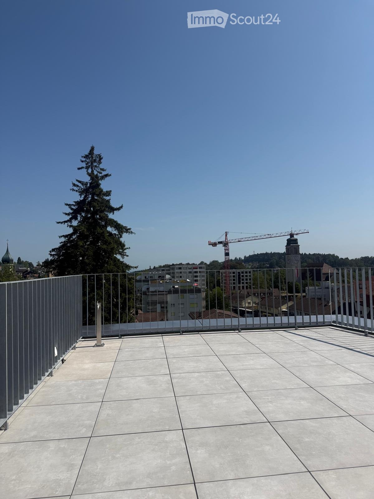 Roof terrace with modern metal railing, tiled floor, and a view of the city skyline, trees, and a construction crane.