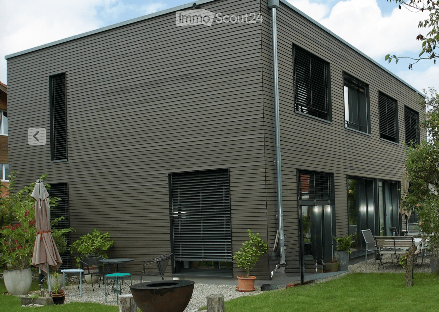 Modern house exterior with grey wooden siding, large windows, and a well-maintained garden area featuring potted plants, outdoor furniture, and a fire pit.