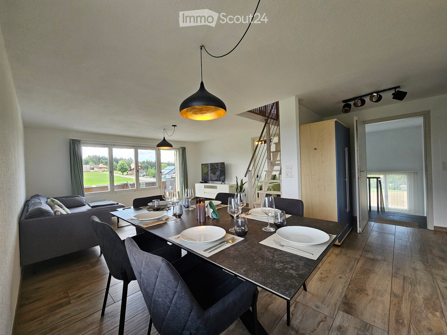 Open-plan living and dining area with a grey sofa, a dark dining table set for six, and a TV on a white stand. The space features wooden flooring, large windows with grey curtains, and modern pendant lights.