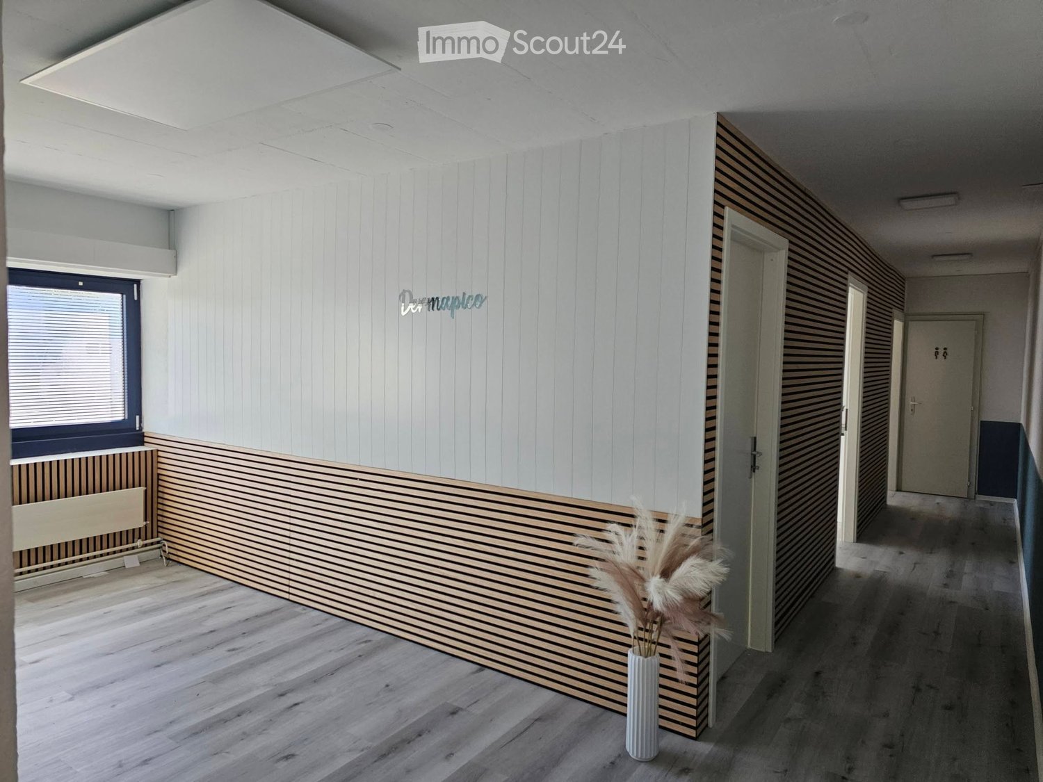 Modern hallway with white paneled walls, wooden flooring, and a decorative vase with dried plants. Features a large window with blinds and multiple doors leading to other rooms.