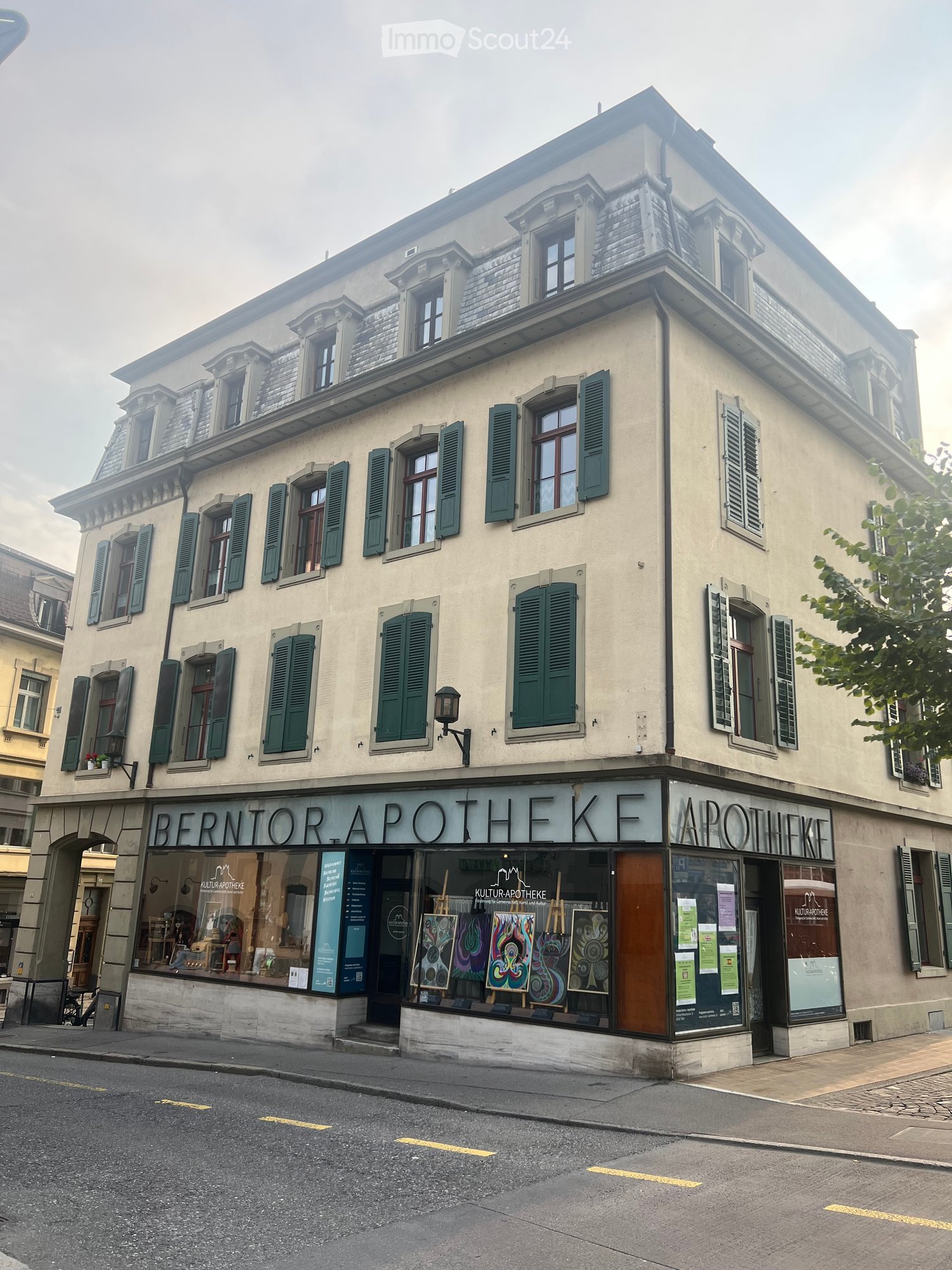 Three-story building with light-colored facade, green shutters, and a storefront labeled "BERNTORE APOTHEKE" with art displays in the window.