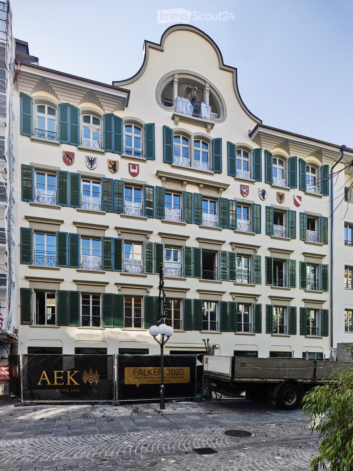 A multi-story building with white facade, green shutters, and multiple windows. The ground floor has a covered entrance area with banners, and the building features decorative emblems and a curved roofline.