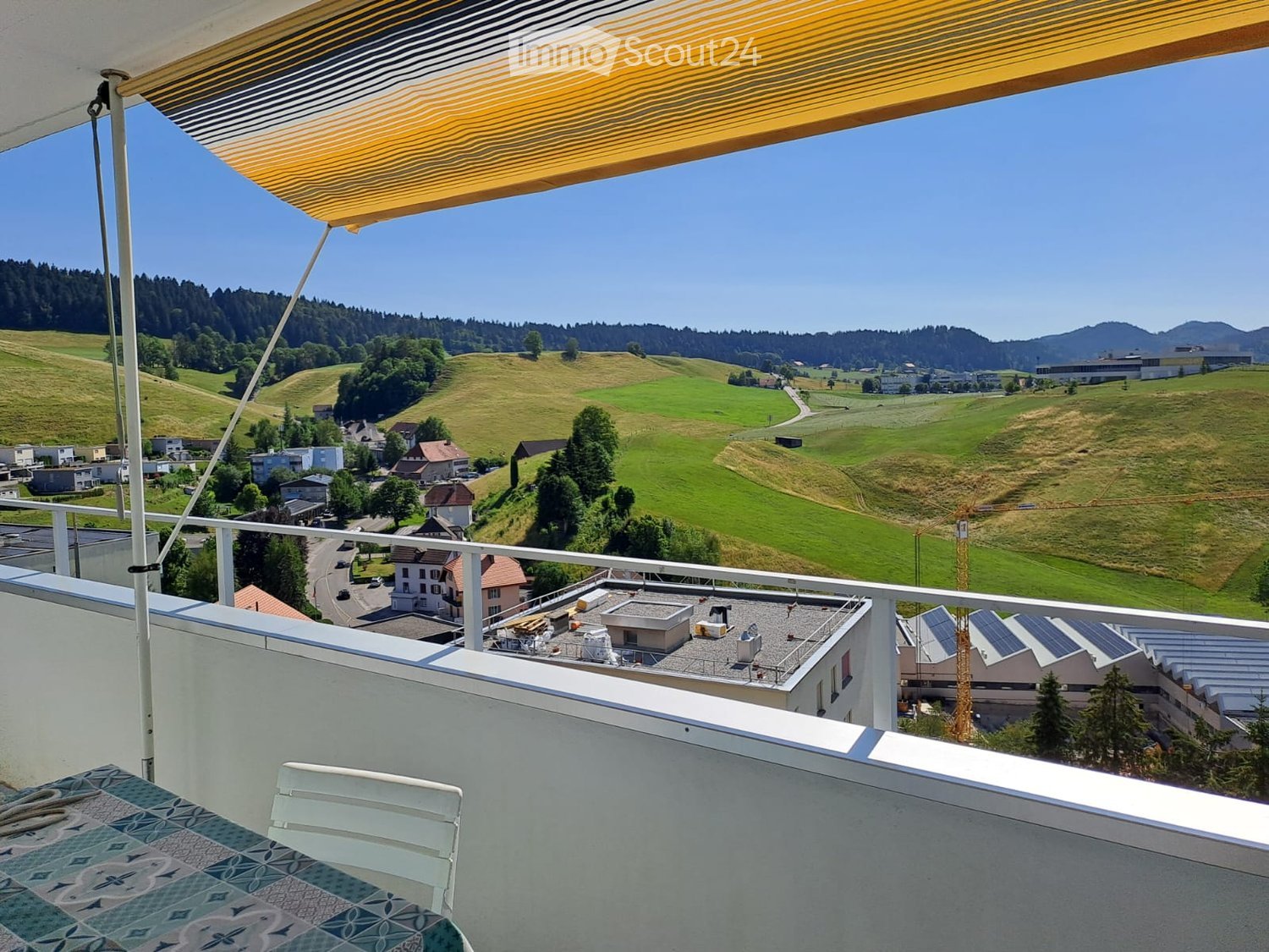 Balcony with a view of a green valley, hills, and scattered houses. Features a white railing, a yellow awning, and a patterned floor.