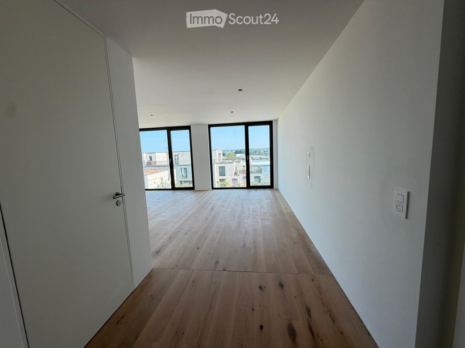 Empty hallway with wooden flooring, white walls, and large windows at the end leading to an outdoor area.