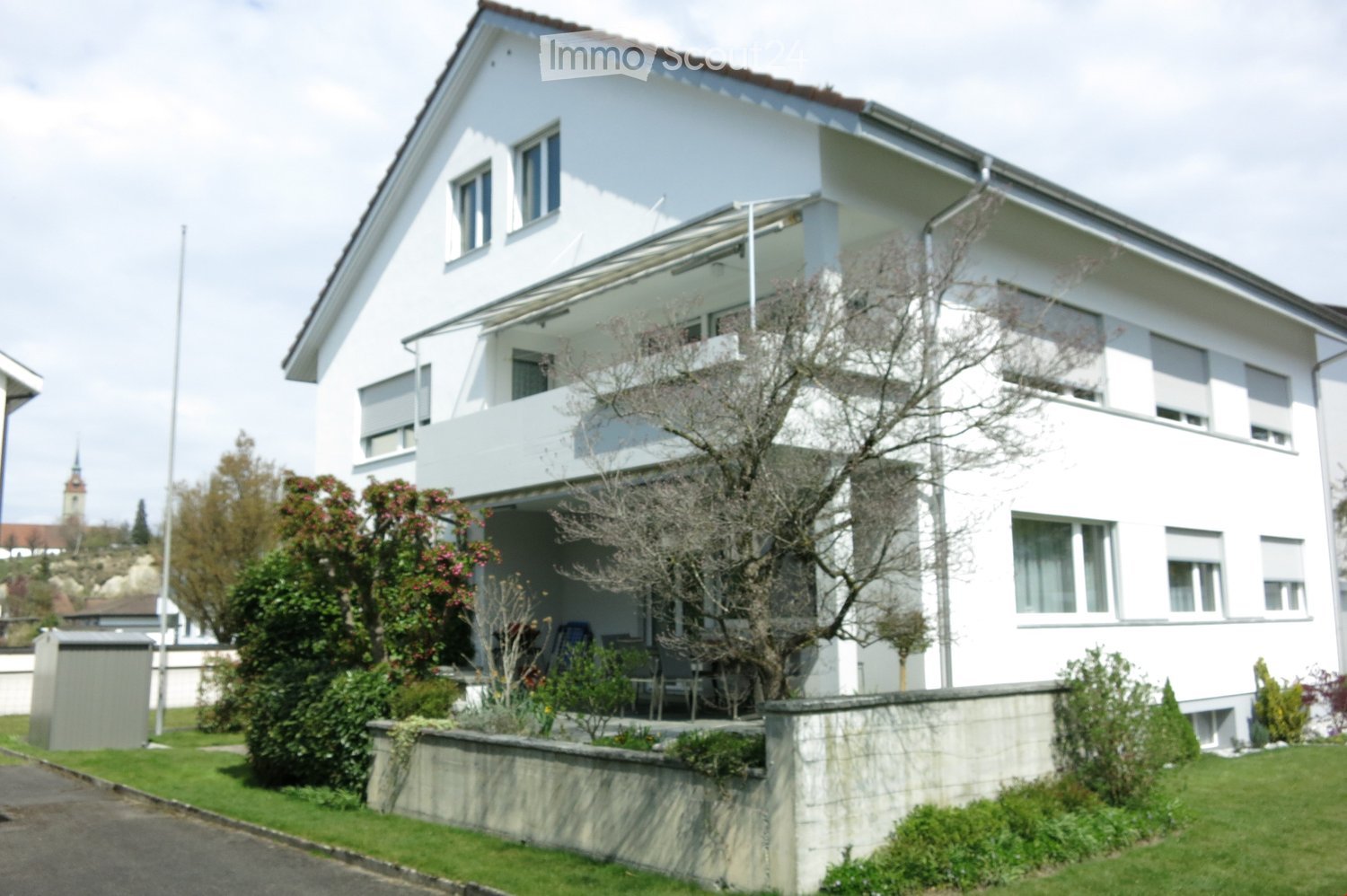 A two-story white house with a modern design, featuring large windows, a balcony, and a well-maintained garden with various plants and shrubs.