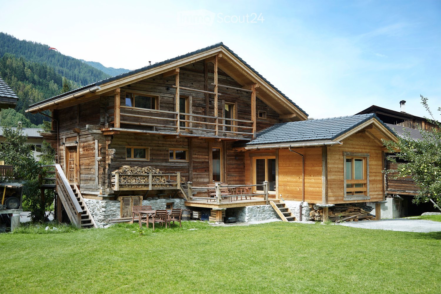 A two-story wooden house with a stone base, featuring a large balcony, wooden deck, and outdoor seating area. The house has multiple windows and a sloped roof, surrounded by a grassy lawn and trees.