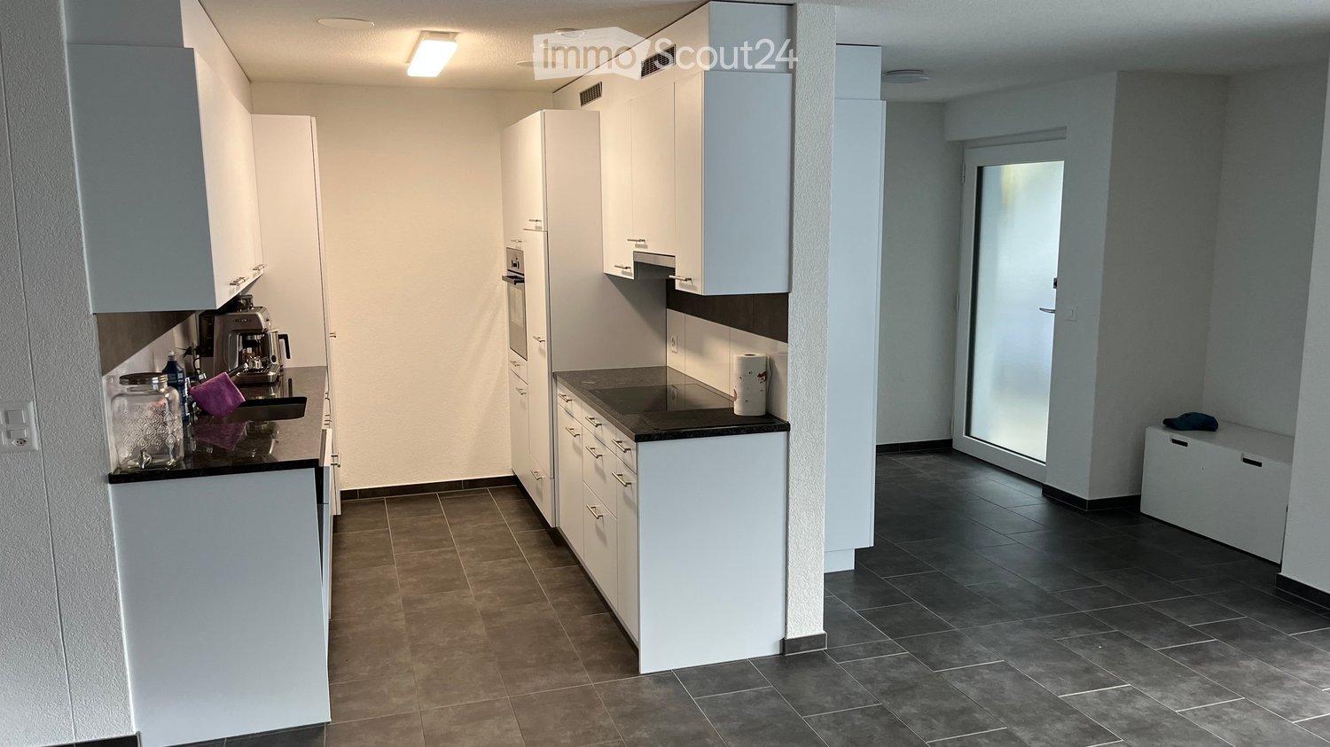 Modern kitchen with white cabinets, dark countertops, and tiled floors. Coffee maker, cup, and jar are visible on the counter.