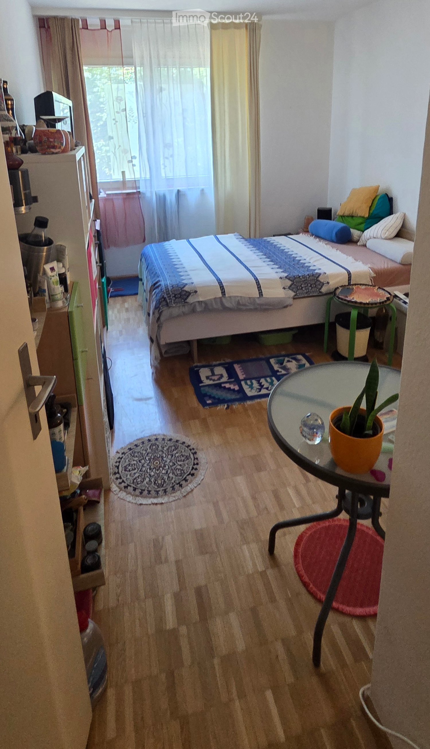Cozy bedroom with wooden flooring, a bed with colorful bedding, a small round table with a potted plant, a stool, and a mirror. The room features a round rug and a small side table with a screen.