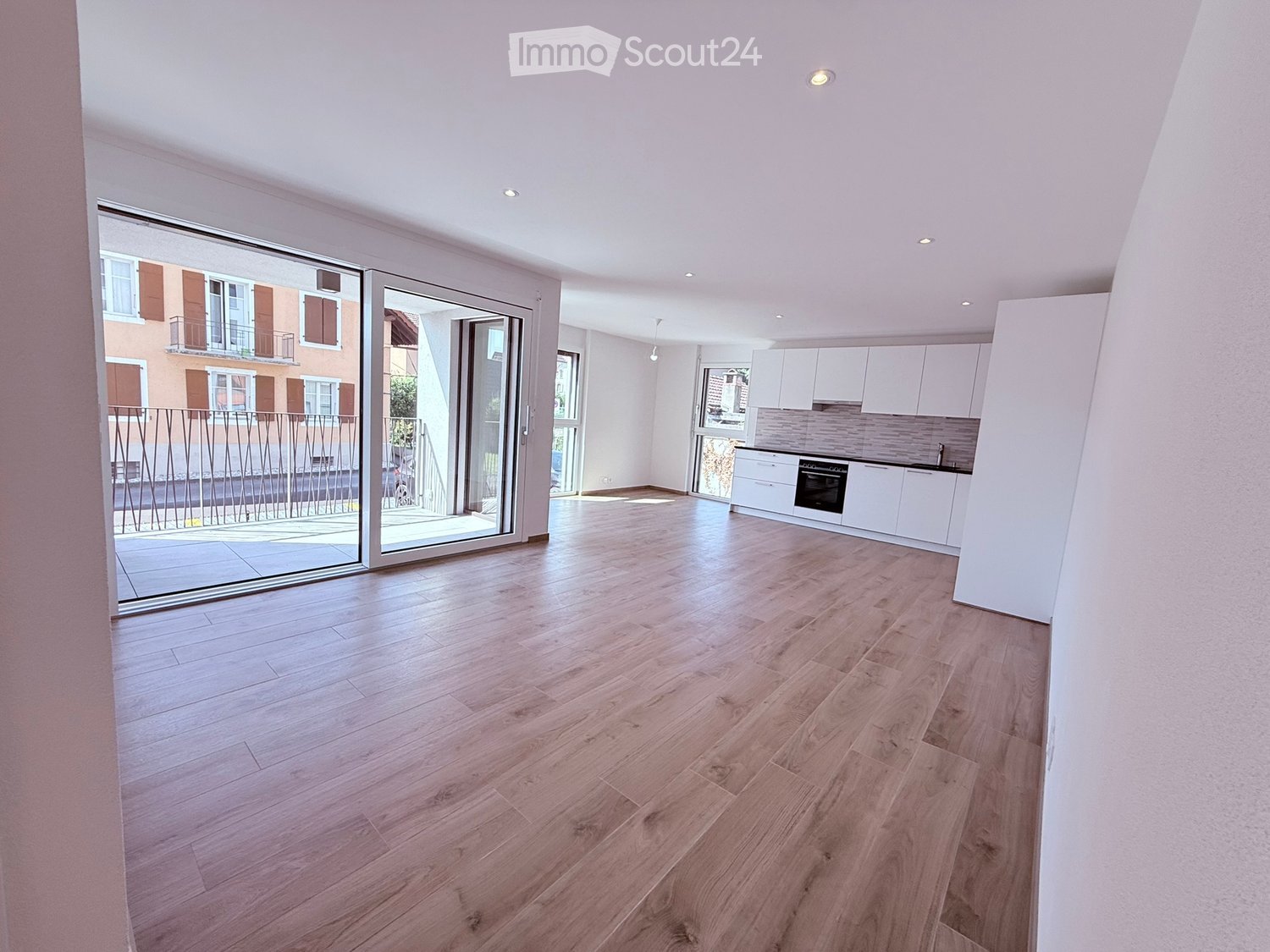 Bright, unfurnished living space with wooden flooring, white walls, recessed lighting, and a large sliding glass door leading to a balcony. Adjacent kitchen area with white cabinets and stainless steel appliances is visible.