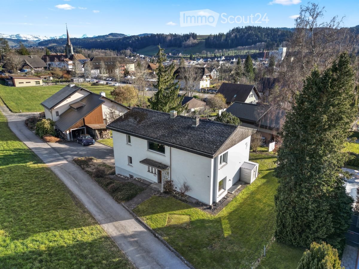 Aerial view of a two-story white house with a dark roof, surrounded by a well-maintained lawn and trees. A driveway leads to a garage with a parked car. The house is situated in a rural area with neighboring houses and a church visible in the background.