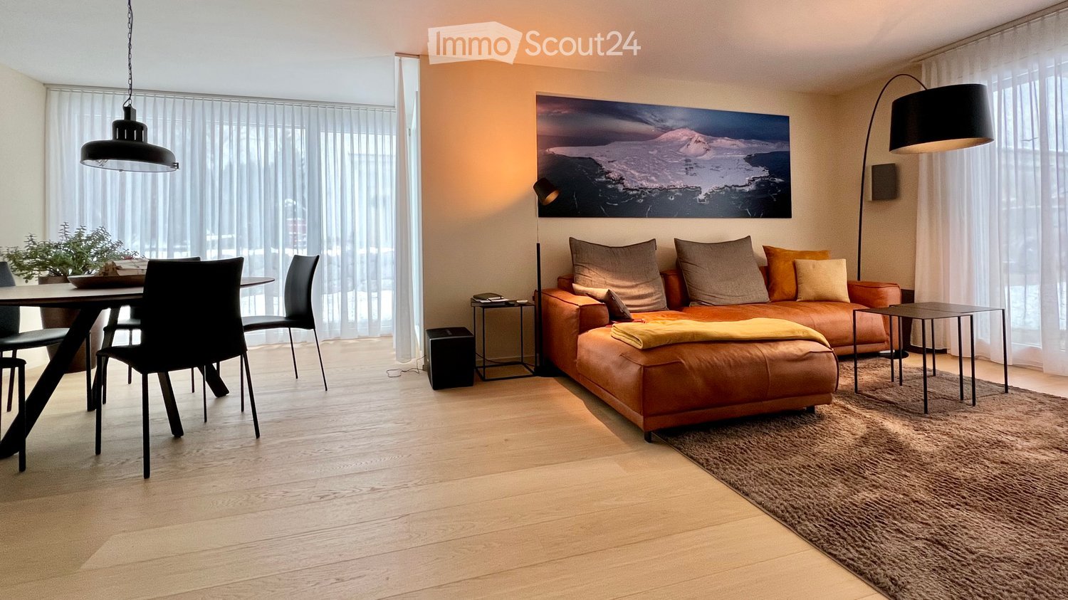 Modern living room with a brown leather sofa, coffee table, dining area with chairs, floor lamp, and large windows with sheer curtains.