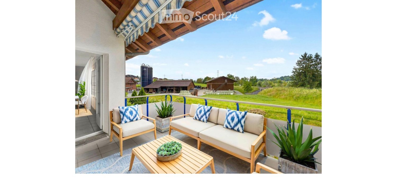 Balcony with a wooden sofa, armchair, coffee table, and potted plants, overlooking a rural landscape with barns and fields.