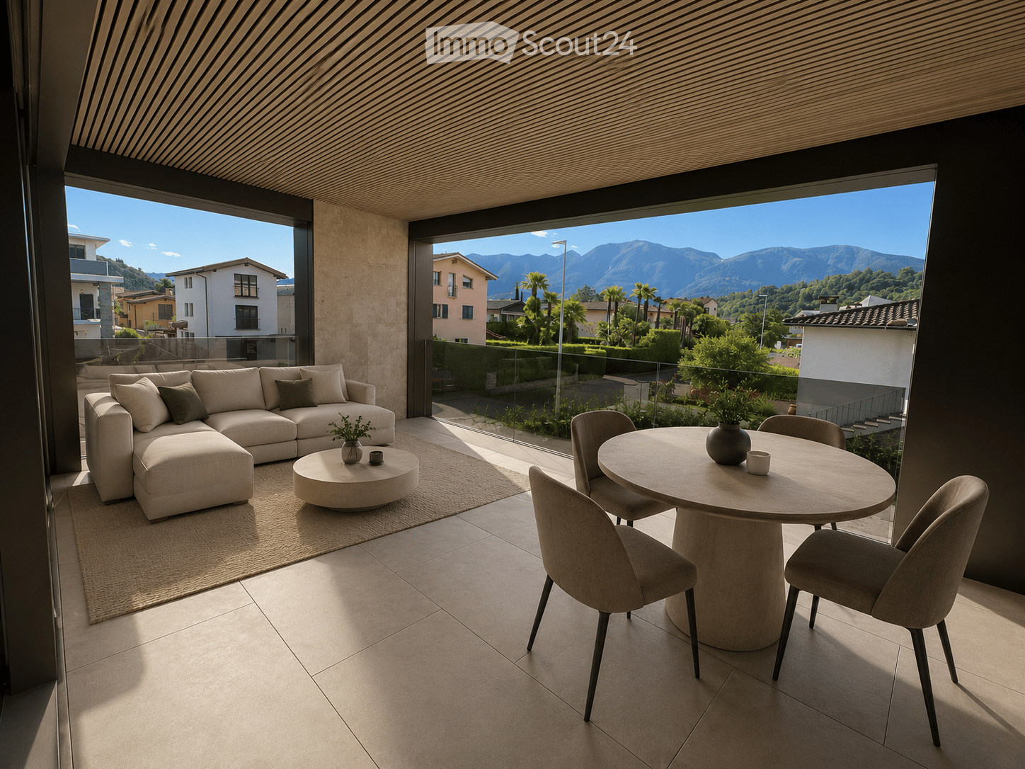 Modern balcony with a panoramic mountain view, featuring a beige sectional sofa, a round coffee table, a dining table with four chairs, and a small vase with plants.