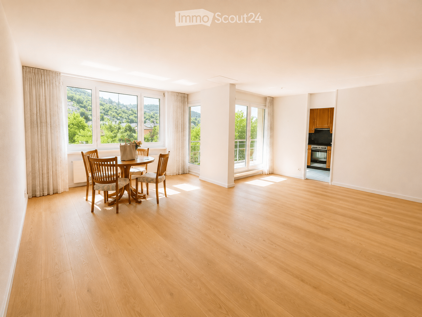 Bright, spacious room with wooden flooring, large windows with curtains, a dining table with chairs, and a handbag with flowers on it. A kitchen area is visible in the background.