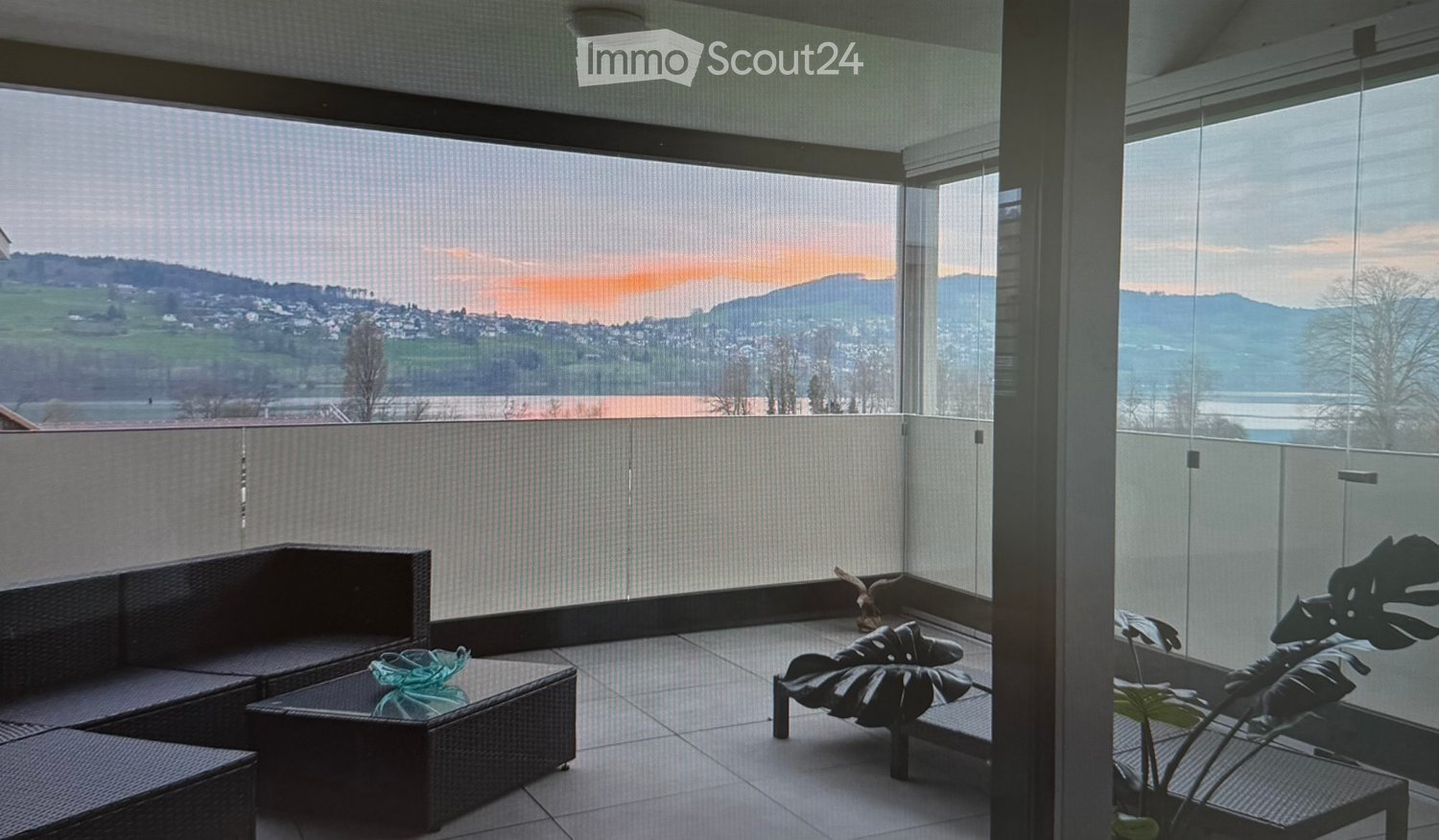Balcony with large windows offering a scenic view, featuring a dark wicker sofa, a small table, a lounge chair, and a leafy plant.