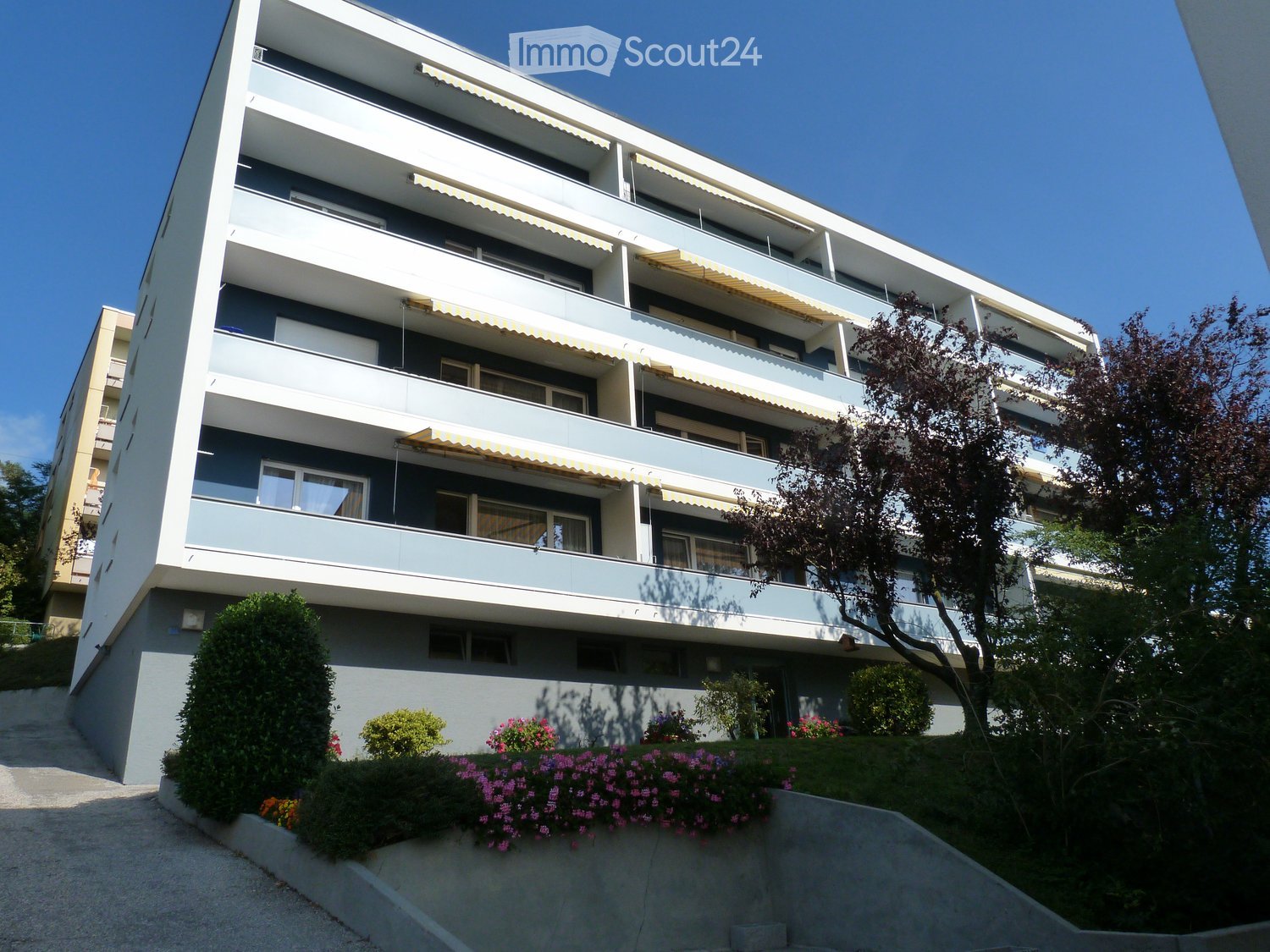 Modern multi-story apartment building with balconies, awnings, and surrounding greenery.
