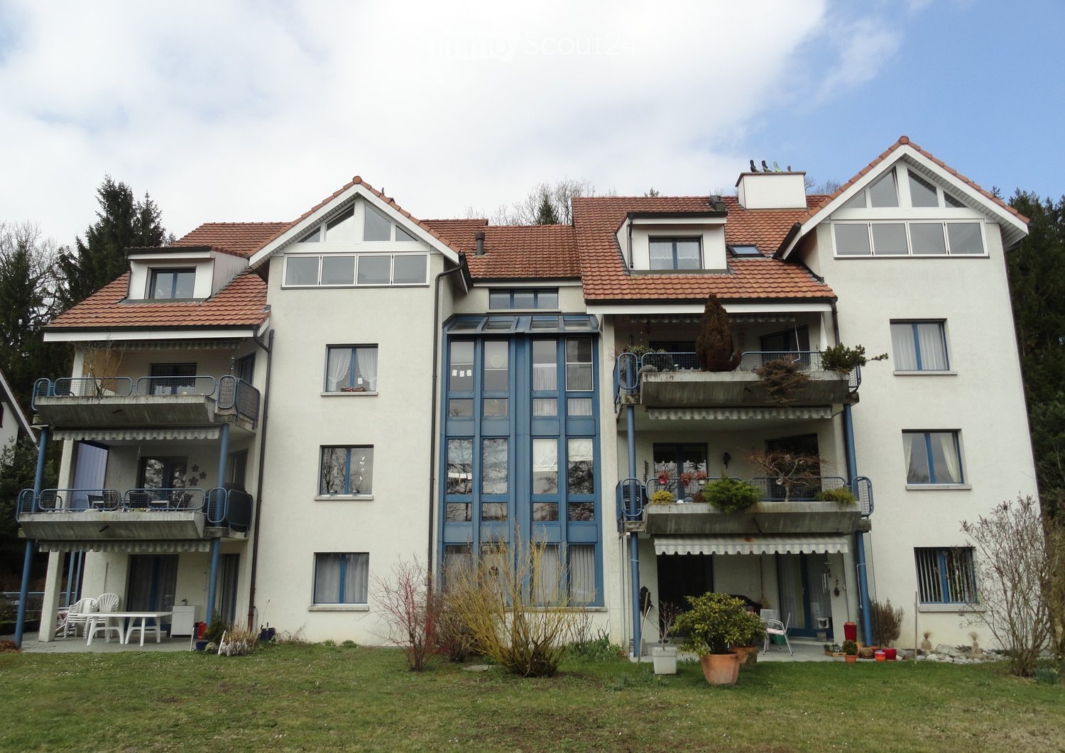 A three-story apartment building with a tiled roof, blue trim, and white exterior. The first floor has balconies with metal railings and white outdoor furniture. There are plants and trees around the building.