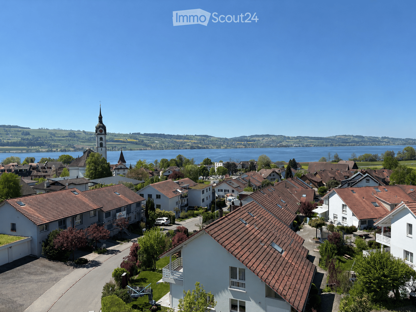 Aerial view of a residential area with multiple houses, red-tiled roofs, and a church tower overlooking a lake in the background.