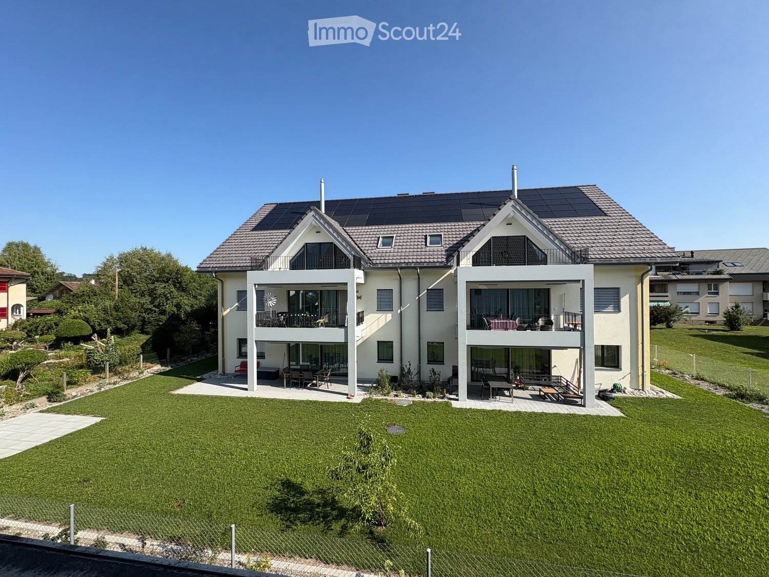 Two-story modern house with solar panels on the roof, large windows, balconies, and a well-maintained lawn.