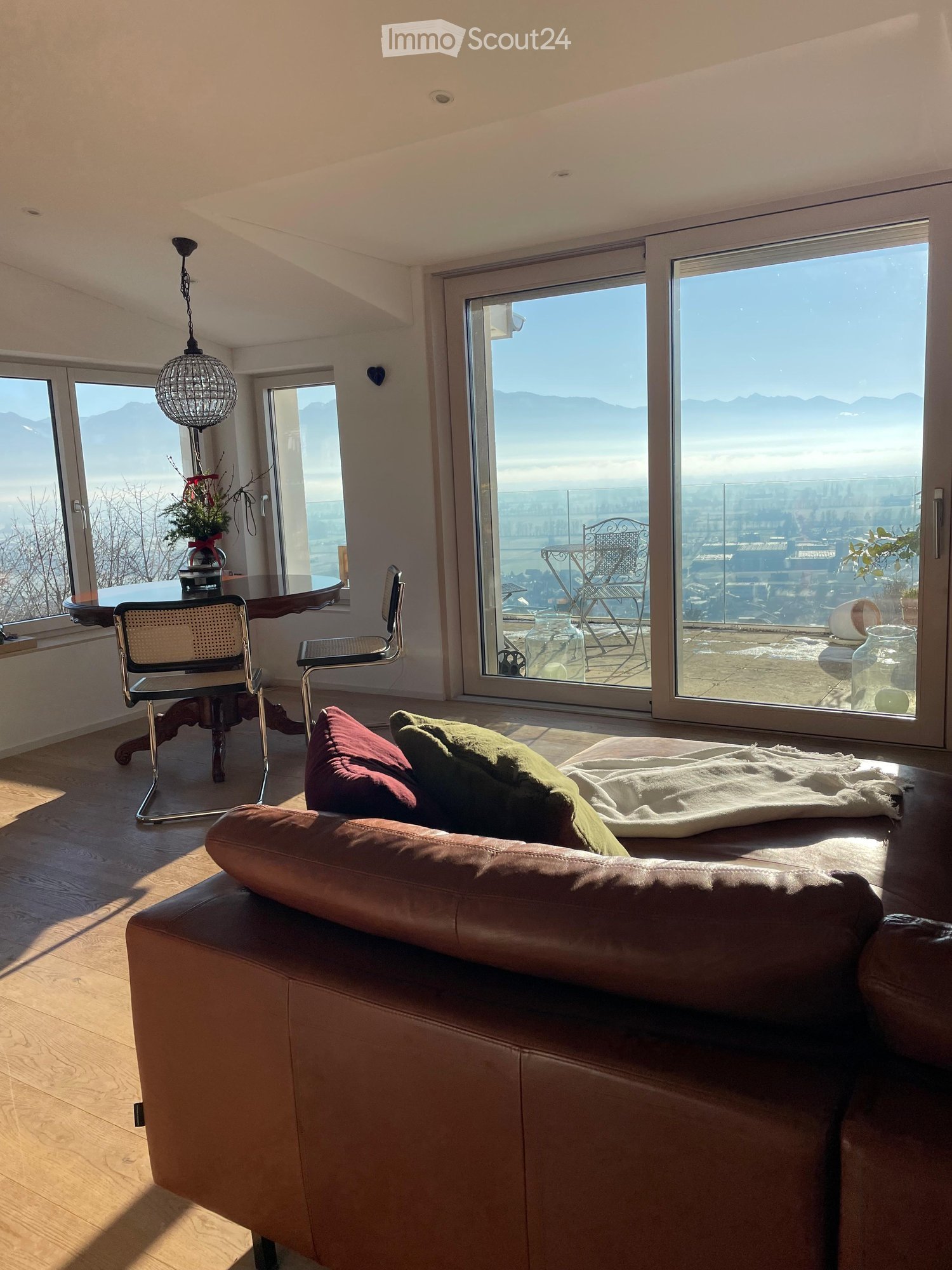 Bright living room with large windows offering a scenic view, featuring a brown leather sofa, throw pillows, a wooden coffee table, and a hanging pendant light.