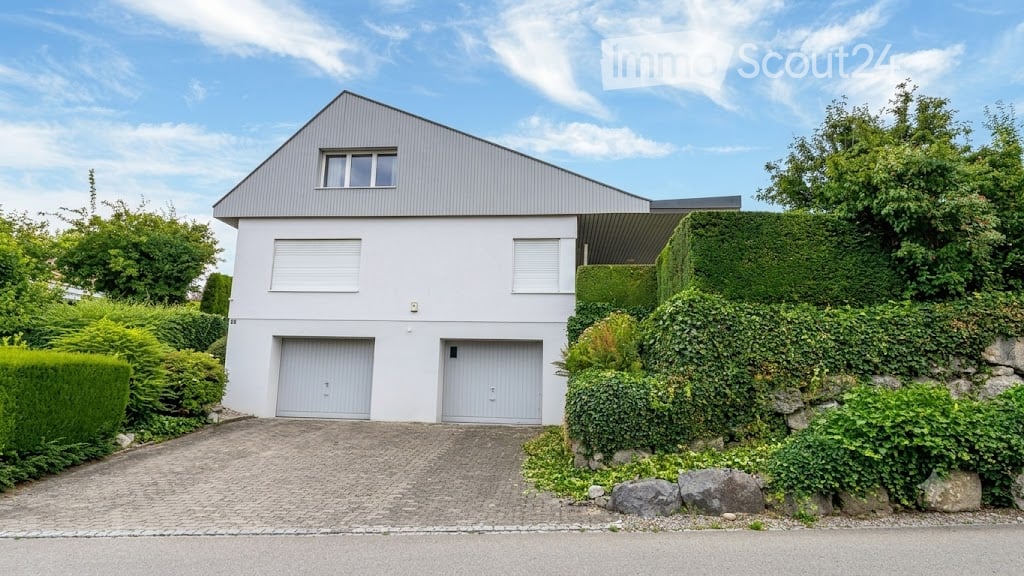 Modern two-story house with white exterior, grey garage doors, and a sloped roof. The house is surrounded by greenery and has a paved driveway leading to the garage.