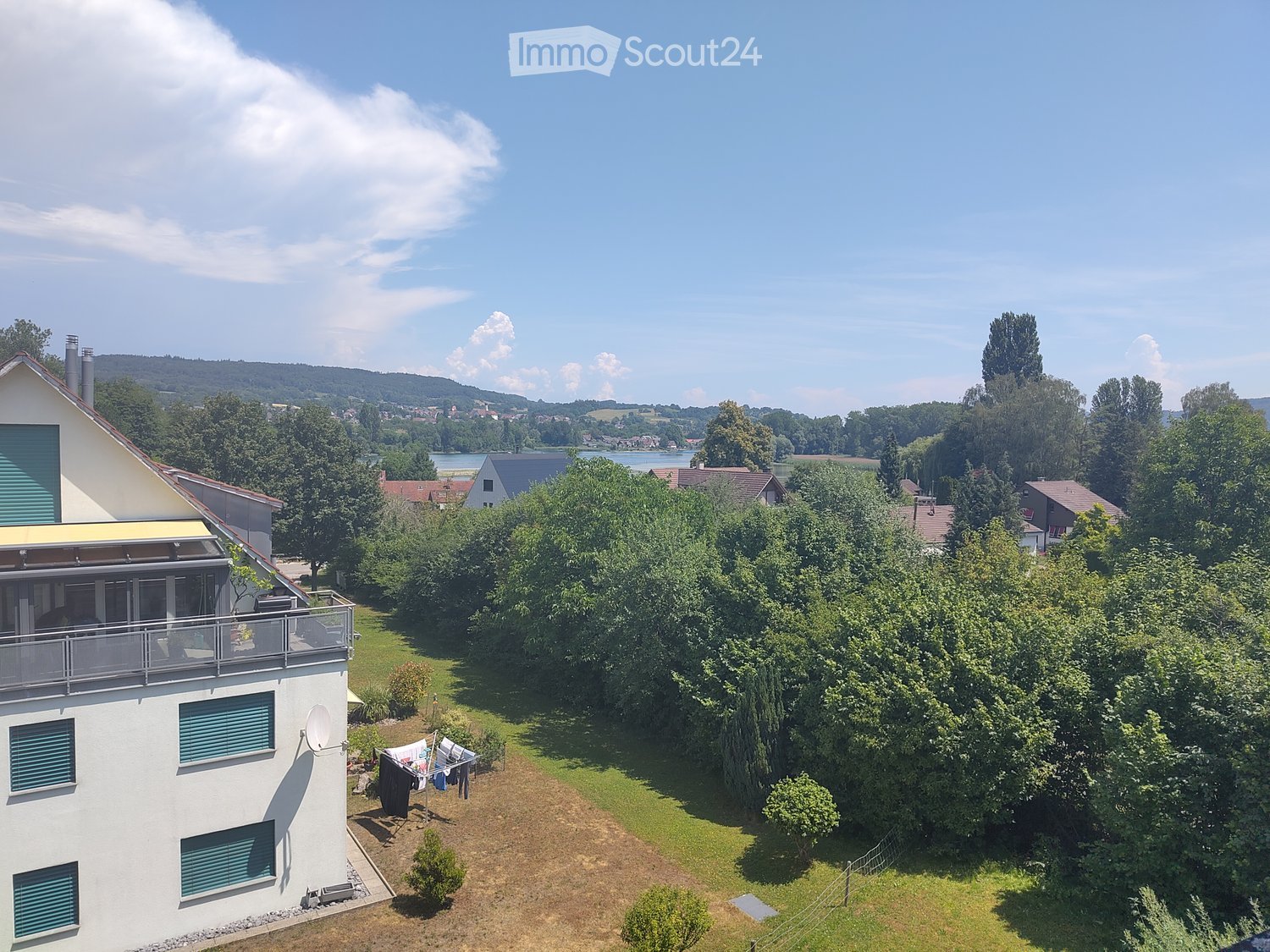 A two-story house with a balcony, surrounded by a well-maintained garden and trees, overlooking a scenic lake and distant hills.