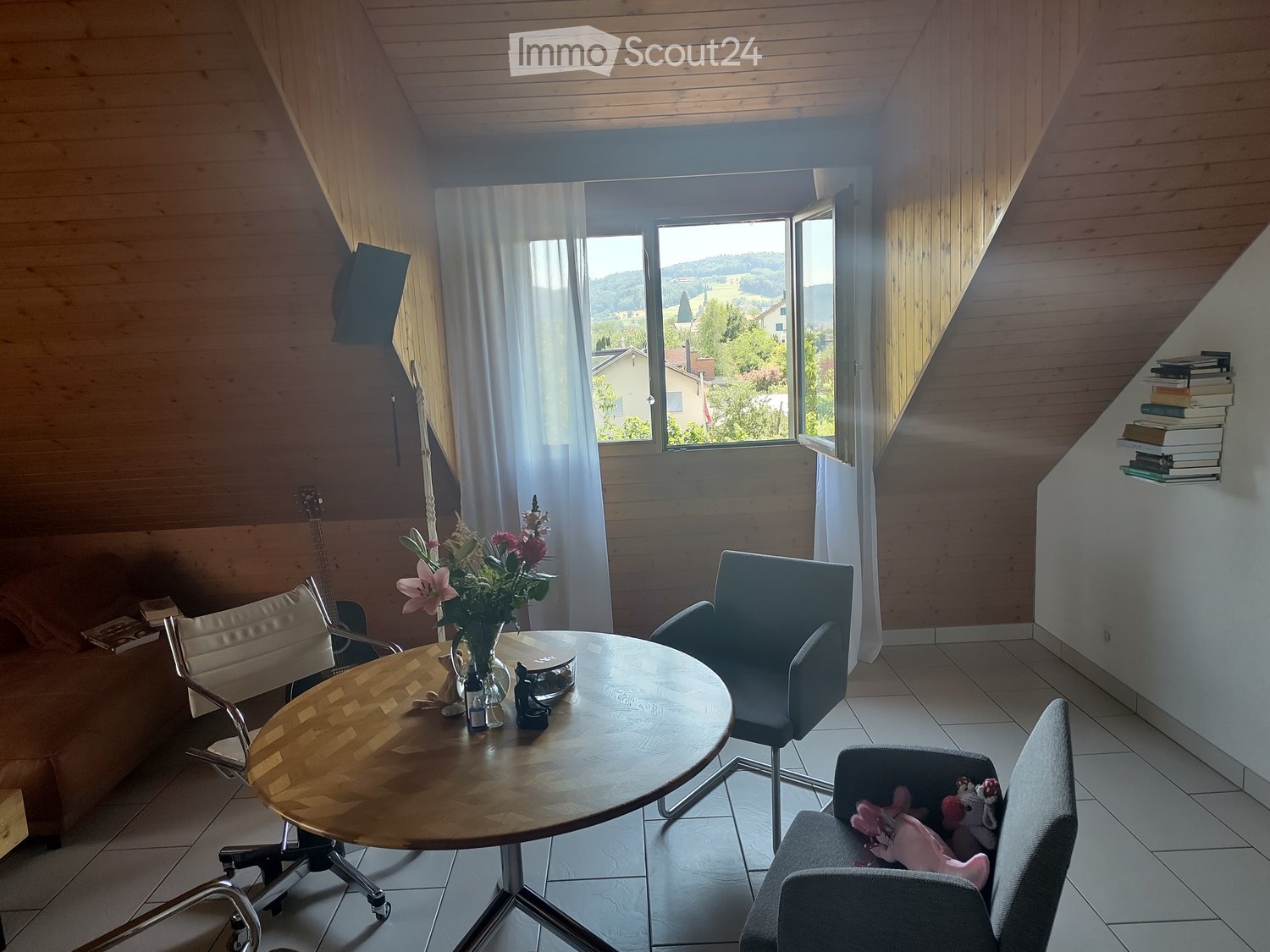Bright living room with wooden walls and ceiling, large window with scenic view, sofa, round wooden table, chairs, guitar, flower vase, bookshelf, and a small stool with stuffed toys.