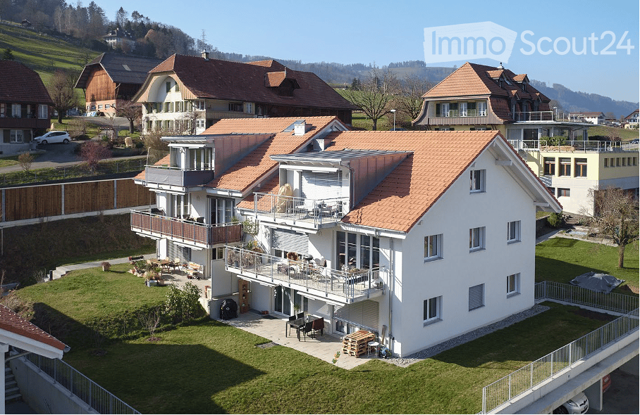 A modern two-story house with a red-tiled roof, white exterior, and multiple balconies. The house is surrounded by a well-maintained lawn and features outdoor seating areas and a patio.
