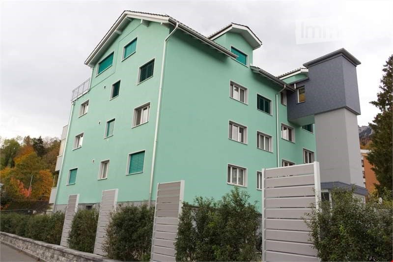 A modern residential building with three floors and several windows. The walls are painted green, and the roof has a triangular shape. The exterior is adorned with multiple white railings on the balcony, and there are bushes and trees on the sides. The bu