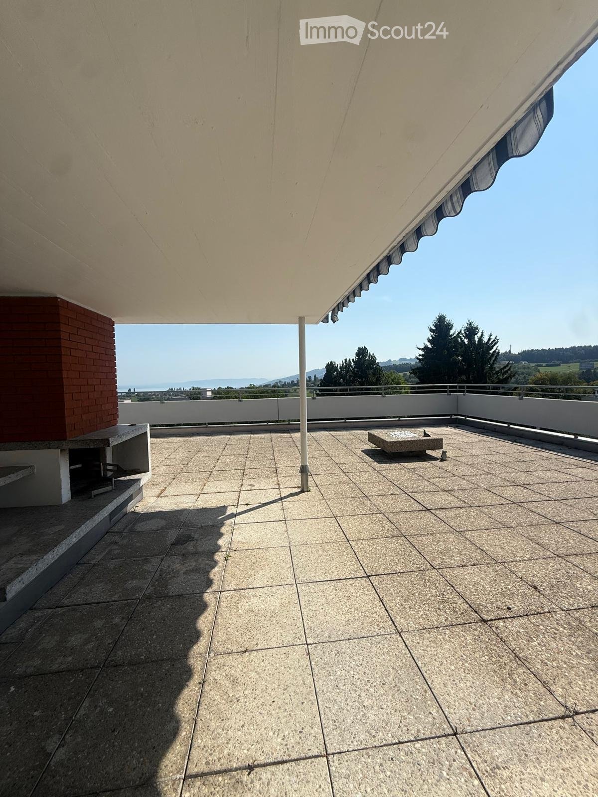 Roof terrace with tiled floor, low white railing, and a small square stone table. The area is covered by a large awning and offers a scenic view of the ocean and surrounding landscape.