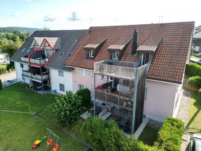 A multi-story residential building with a red-tiled roof, multiple balconies, and a modern glass balcony structure. The building is surrounded by a well-maintained lawn and trees.