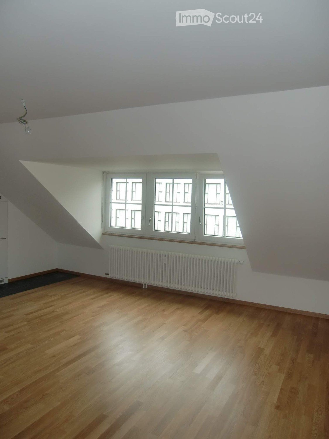 Empty room with wooden flooring, white walls, and large windows with white frames. A radiator is visible below the windows.