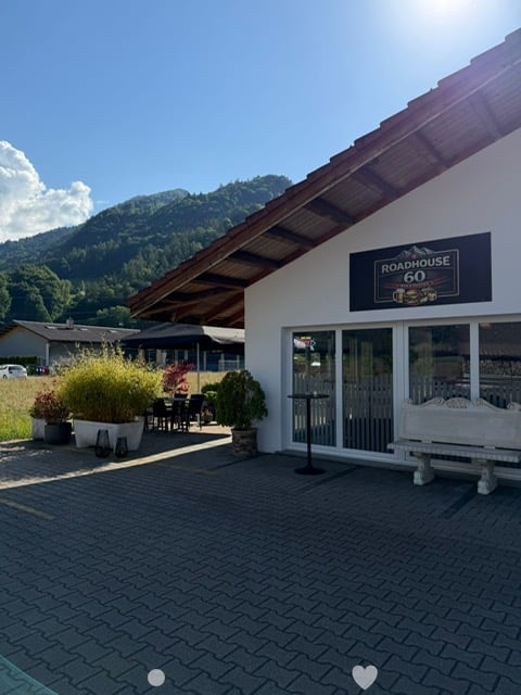 Outdoor seating area of a restaurant with a sign reading "ROADHOUSE 60" on the building. There are potted plants, a small table with chairs, and a bench near the entrance.