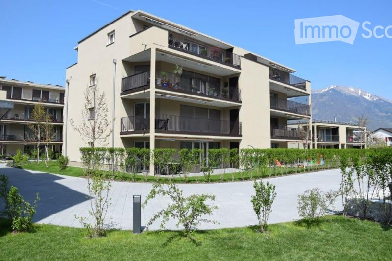 Modern multi-story apartment building with balconies, surrounded by greenery and a clear view of mountains in the background.