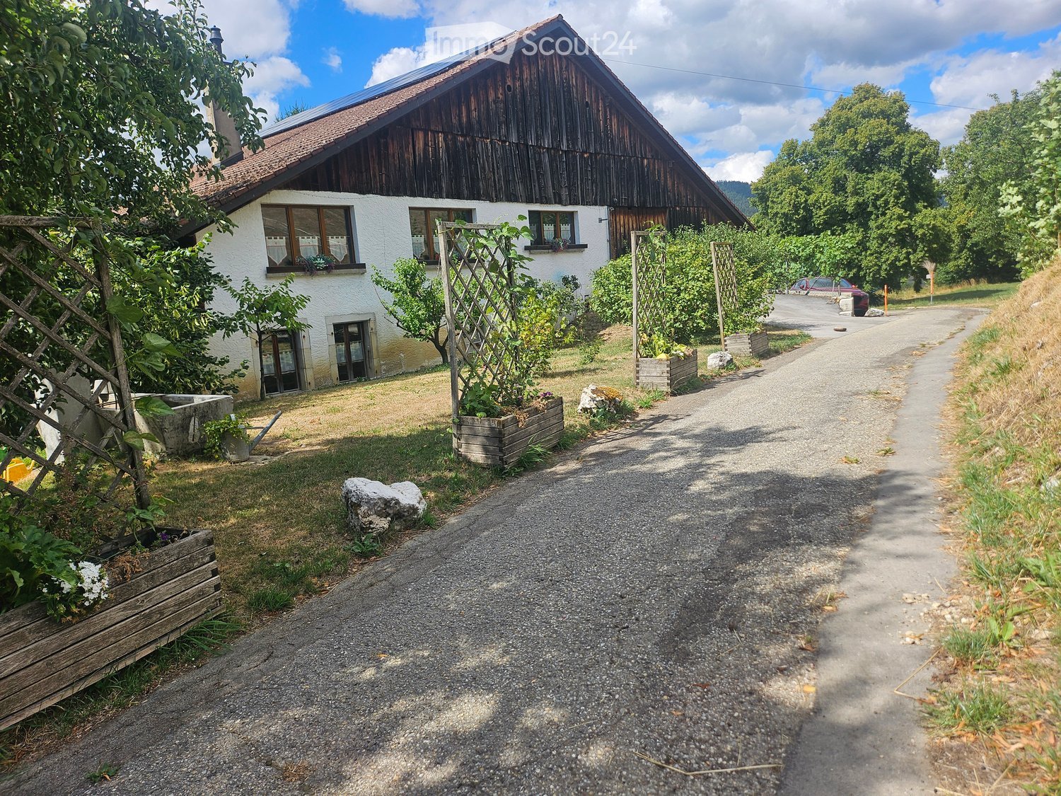 A two-story house with a wooden exterior and white walls, surrounded by greenery and a gravel driveway.