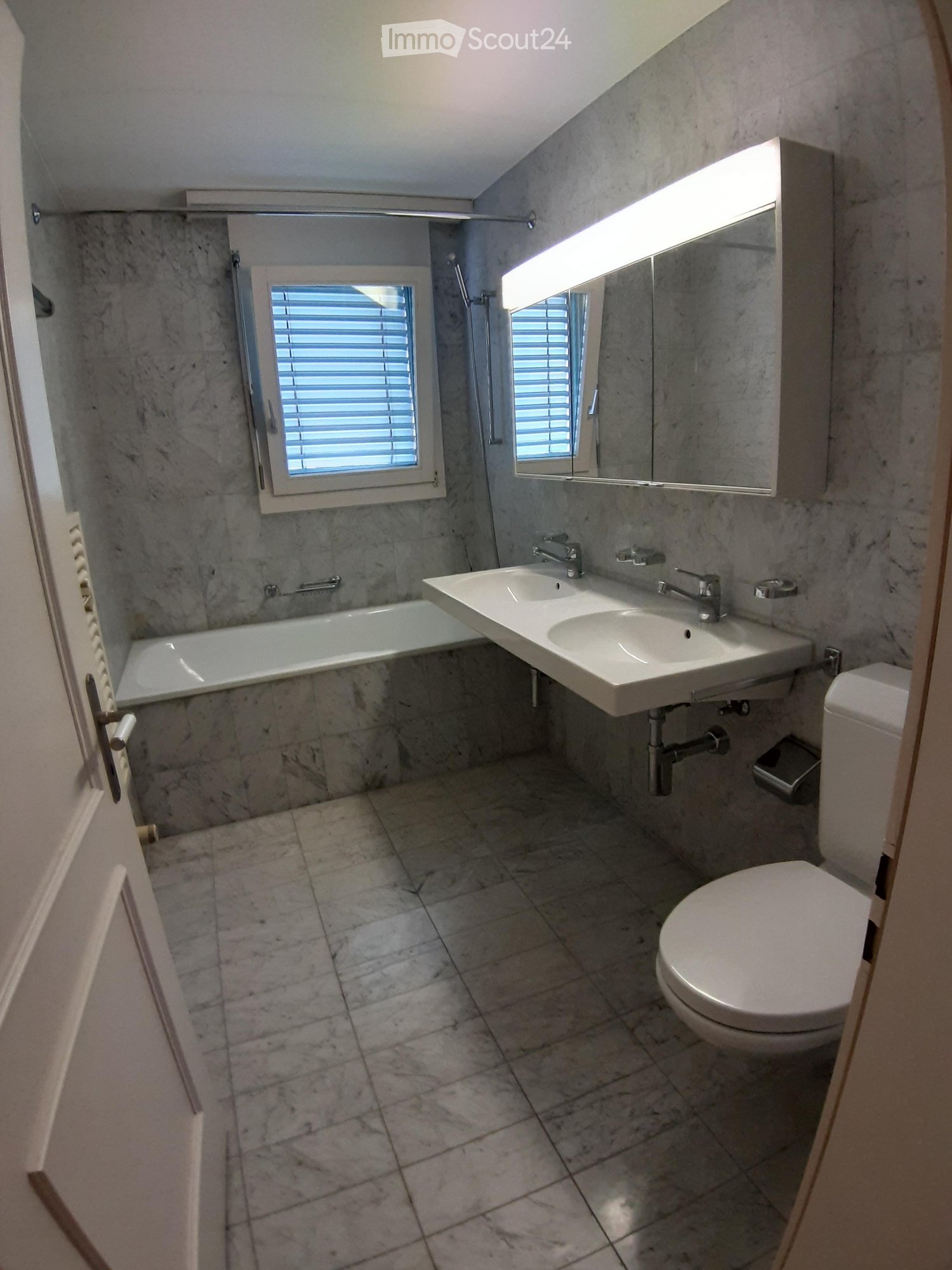 Modern bathroom with marble tiles, a bathtub, double sink, toilet, and a large mirror with built-in lighting.