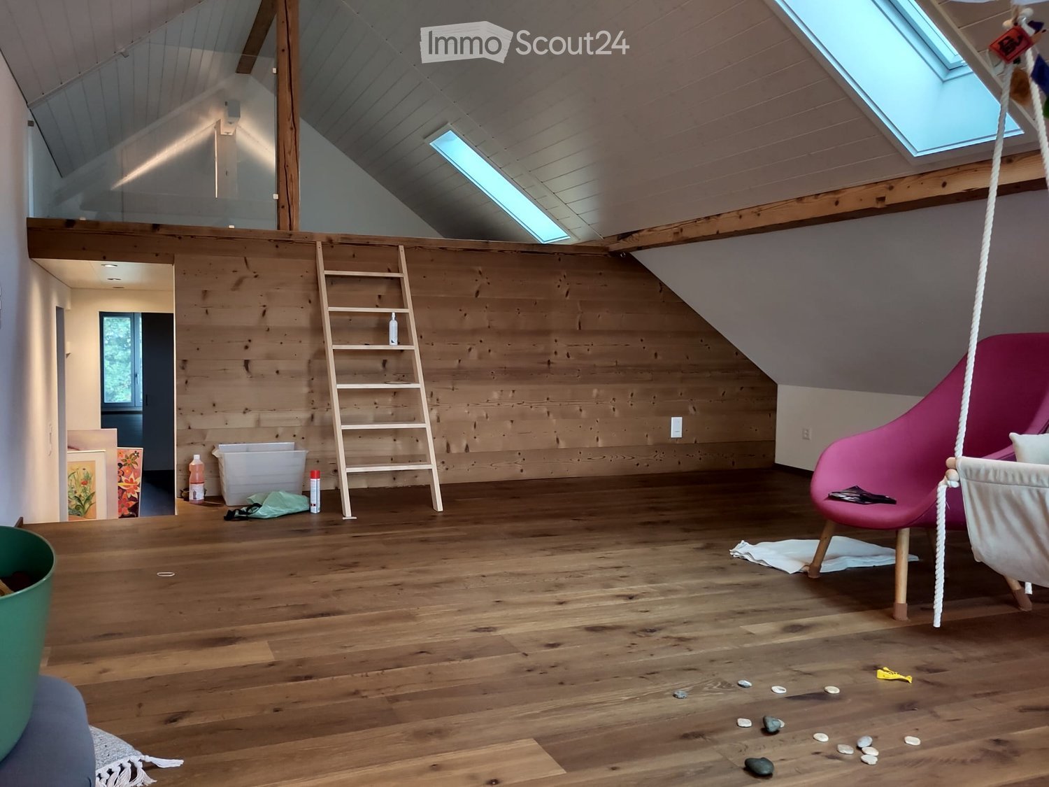 Loft-style room with wooden floors, exposed wooden beams, skylights, a pink chair, a ladder, and a wicker hanging chair.
