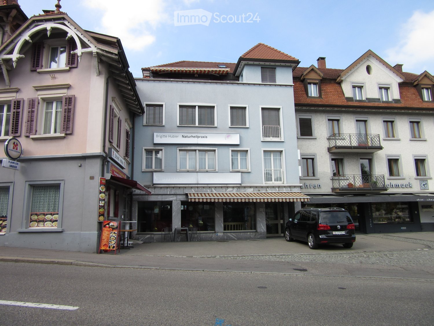 A row of multi-story buildings with shops and apartments, featuring awnings, windows, and a parked car.