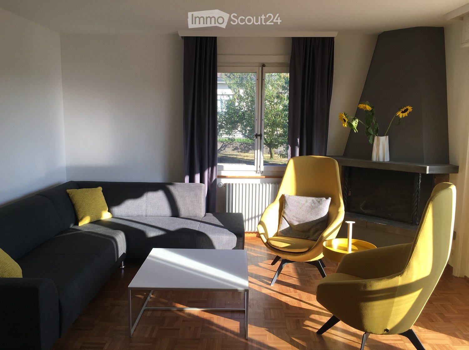 Modern living room with a grey sectional sofa, yellow chairs, a white coffee table, and a fireplace with a vase of sunflowers on the mantel.