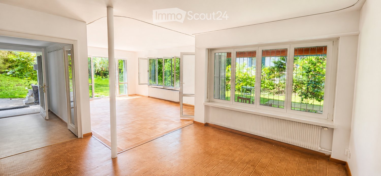 Bright, empty room with wooden flooring, large windows, and an open doorway leading to a garden area.