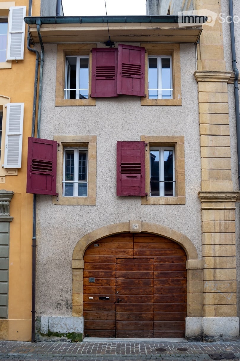 Facade of a two-story building with a wooden garage door, multiple windows with red shutters, and a textured beige wall.