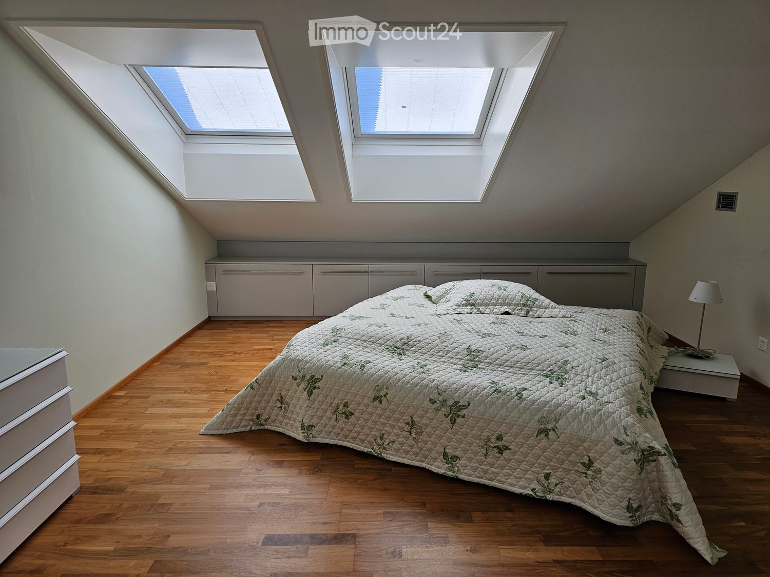 bedroom with wooden floor, double bed with quilt, two windows with skylights, nightstand, lamp, drawers