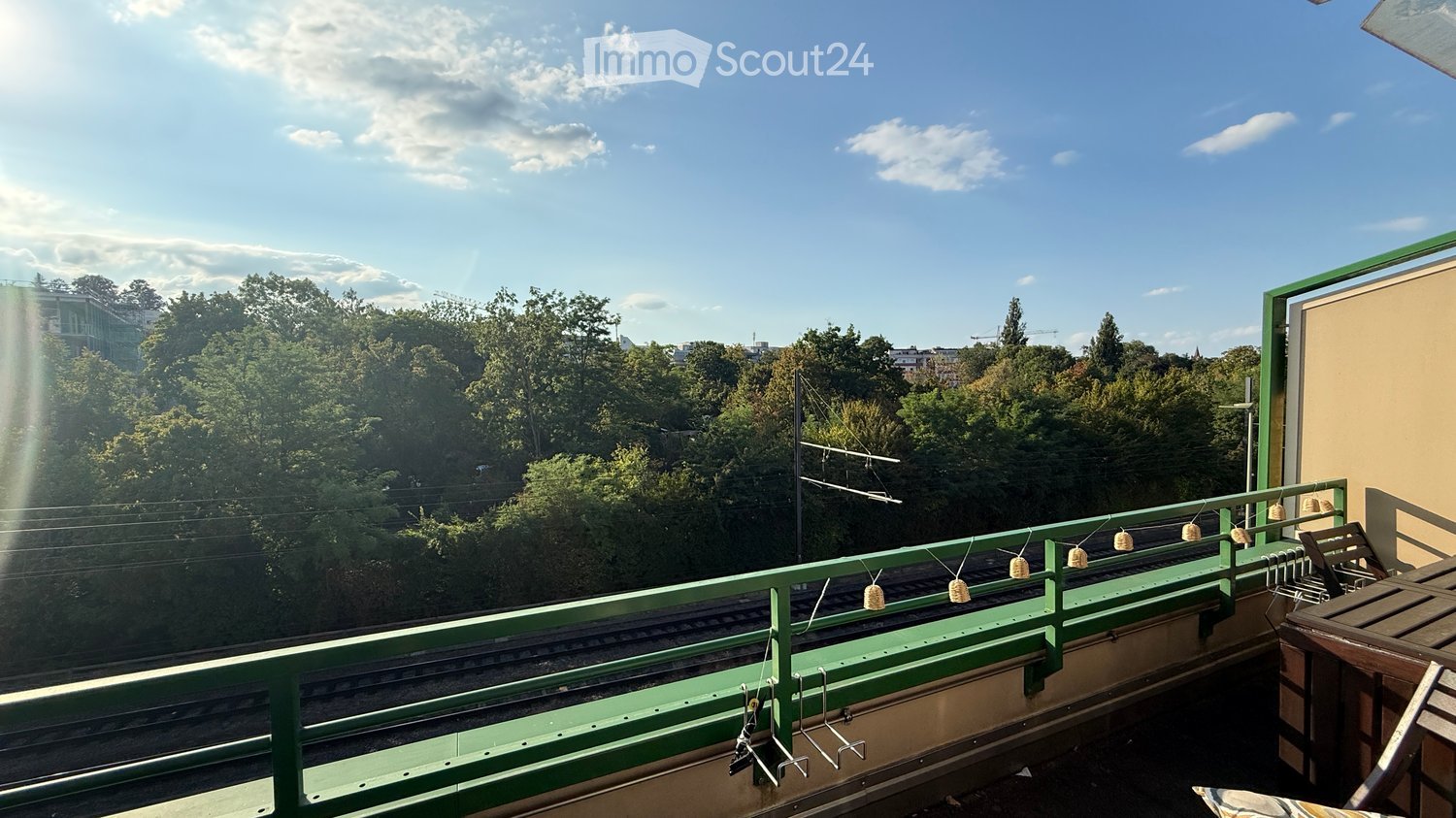 Balcony with green railing, wooden table, and chairs, overlooking a tree-filled area with a cityscape in the background.